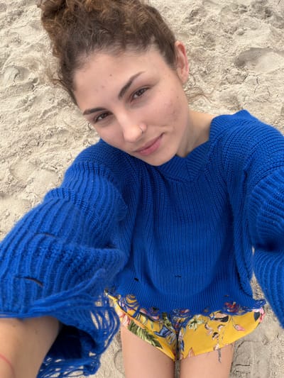 A young woman is lying on a sandy beach, possibly in Kiryat Yam, Israel. She is wearing a bright blue, long-sleeved knit sweater with fringed edges and a yellow floral-patterned dress or swimsuit. Her head is turned towards the camera, and she is gazing directly at it with a slightly tilted face. She has curly light brown hair and fair skin. The background consists of sand, with subtle indentations suggesting footprints or disturbances. The overall impression is a casual, personal photograph taken at eye level.