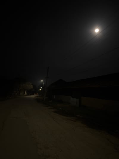 The image depicts a nighttime scene in Maslarevo, Bulgaria. A dirt or gravel road, marked with cracks and visible wet patches, extends from the foreground into the distance, gently curving left. On the left side of the road, sparse, bare trees or bushes are silhouetted against the dark. Further down this path, a second, fainter streetlight emits an orange glow. On the right side of the road, a utility pole with a bright white-yellow streetlight stands prominently, illuminating the immediate road surface and the facade of an adjacent low-slung building. This building features a dark, sloped roof and a lighter-colored wall or fence segment with a light-toned gate or door. Overhead, power lines traverse the dark sky, beneath a prominent, bright moon positioned in the upper right quadrant, appearing slightly diffused by atmospheric conditions. No individuals, animals, or vehicles are present.