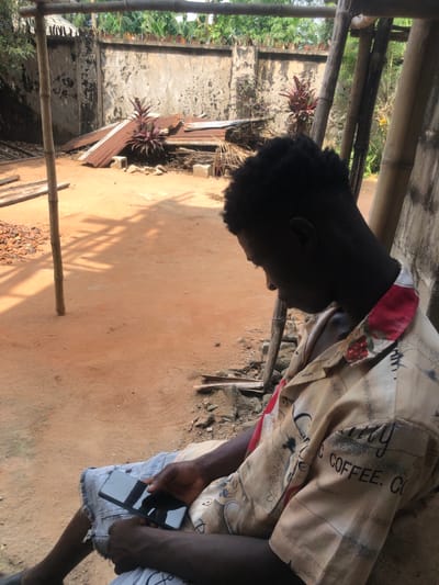 An outdoor scene in Abagana, Nigeria, depicts a male individual seated on the right side of the frame, facing downwards and left. He possesses dark skin and short, dark, curly hair. He wears a light-colored, short-sleeved collared shirt patterned with coffee-related motifs and red designs on the collar, along with light-colored, distressed shorts. The individual holds a black smartphone with a reflective screen in his left hand, resting on his lap, and appears engaged with the device.
The ground consists of exposed red-orange soil, illuminated by strong sunlight that casts shadows, including one from a vertical bamboo pole located in the left mid-ground. Near the base of this pole, small reddish-brown rocks or debris are present. The background features a weathered, light-colored concrete or stucco wall with staining and an irregular top edge. Behind and above the wall, green foliage is visible. Against the wall, a collection of objects includes several pieces of corrugated metal, some rusted, piles of dried plant material resembling palm fronds, wooden planks, and two distinct plants with reddish-purple leaves. Further bamboo-like structures are visible on the far right.