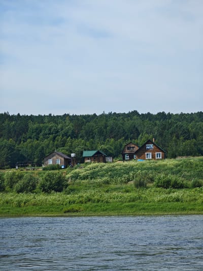 A wide, slightly rippled river flows in the foreground, reflecting the blue sky and white clouds above. The riverbank is lined with lush green grass and dense vegetation. On the elevated bank, a small cluster of wooden houses is visible. The largest house, in the center, is made of dark wood and has white-framed windows. A blue tarp is visible in front of it. To its left, a smaller, lighter-colored wooden house with white-framed windows stands. Further to the left, another small building with a green roof is nestled amongst the greenery. Behind the houses, a dense forest of green trees stretches across the horizon. The overall scene depicts a tranquil, rural landscape. The location is identified as Porog, Russia. There are no people visible in the image.