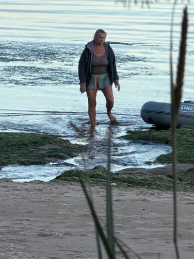 An adult woman is standing in shallow water near the shore of Fort Krasnaya Gorka, Russia. She is wearing a dark jacket over a brown top and green shorts with an orange stripe. The water is calm and reflects the sky, which appears overcast. To the right of the woman, a portion of an inflatable boat with the word "LINC" visible on its side is partially submerged in the water. The foreground shows a sandy beach with some green vegetation and rocks. The overall scene suggests a natural outdoor environment.