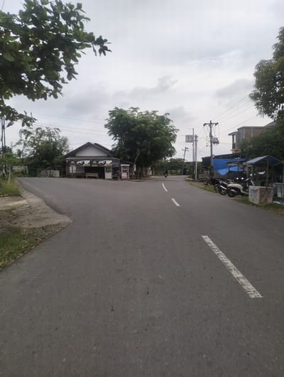 The image shows a street scene in Muliengmanyang, Indonesia, on an overcast day. A paved road curves from the foreground towards a small cluster of buildings on the left and right sides of the road. The building on the left appears to be a small shop with a peaked roof and a sign that reads "RIMA". Several motorcycles are parked in front of structures on the right side of the road, one with a blue tarp roof. A single motorcycle is visible in the distance on the road, heading away from the viewer. Trees and utility poles are scattered throughout the scene. The sky is heavily clouded.