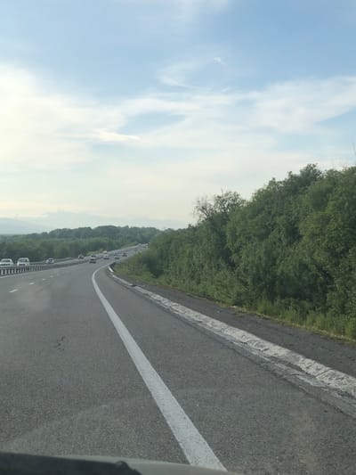 The image shows a highway with traffic, bordered by lush green trees on the right and a protective barrier on the left. The sky is partly cloudy with a bright, light blue hue. In the distance, a mountain range is faintly visible under the hazy sky. Cars are seen driving on the highway, with a few in the foreground and more in the distance, suggesting a moderate flow of traffic. The road surface is asphalt, marked with white lane dividers. The scene depicts a typical highway environment, likely in a rural or semi-rural area. There are no people visible in the image. The presence of a highway suggests it is a transportation corridor. The vegetation indicates a temperate climate. There is no specific information in the image to confirm the location in Svetly, Russia.