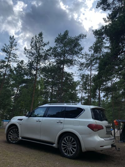 This media file is a photograph of a white SUV parked on a dirt path in a forest. The car is positioned diagonally in the frame, with its rear to the right. Pine trees with dark green needles surround the scene, and the ground is covered in dry grass and pine needles. The sky above is cloudy and overcast, suggesting a possibly rainy or stormy day.
In the background, partially visible to the right of the SUV, are two tents and what appears to be camping equipment, indicating that the setting is a campsite. A license plate is visible on the rear of the car, but the text on it is not legible. The overall mood of the image is serene and rustic, conveying a sense of being in nature. The lighting is diffused due to the cloudy sky, and there are no people visible in the frame. The photo appears to have been taken during daylight hours, likely in the afternoon or evening given the subdued light.