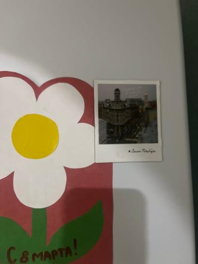The image displays a portion of a wall decorated with a large white and red flower illustration. Attached to the wall, to the right of the flower, is a small polaroid-style photograph featuring a cityscape. The photograph shows buildings with architectural details and a dome, suggesting an urban European setting. Below the cityscape in the photograph, handwritten text in Cyrillic script is visible, which appears to read "С любовью - Петербург" (With love - Petersburg). The bottom portion of the flower illustration shows the text "С 8 МАРТА!" (Happy 8th of March!) also in Cyrillic. The overall context suggests a celebratory decoration, possibly for International Women's Day, with a personal touch referencing St. Petersburg. The image does not contain any information to suggest the location is Gorodets, Russia.