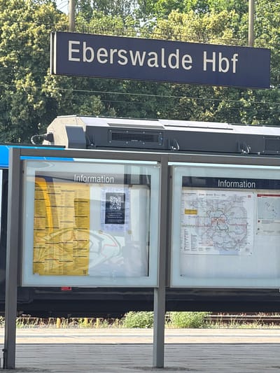 This is a vertical, eye-level shot of a train station sign and information boards. The sign, in dark blue with white lettering, clearly reads "Eberswalde Hbf," indicating the Eberswalde Hauptbahnhof (main station) in Germany. To the right of the sign are two large, glass-enclosed information boards. The upper board displays a map, likely a public transportation route map, with various lines and stations marked. The lower board also contains maps and schedules, with a QR code visible in the upper right corner, suggesting a digital information link. The text "Information" is prominently displayed on both boards. The scene is outdoors, with lush green trees visible in the background, suggesting a sunny day. The lighting indicates it is daytime. The train tracks and a wooden platform are visible to the right of the information boards. There are no people visible in the image. The overall impression is that of a well-maintained public transport hub on a pleasant day.