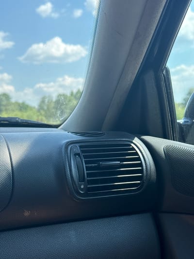 The image shows the interior of a car, with a focus on the dashboard and a portion of the windshield. The dashboard features a black textured surface and an air vent with adjustable slats. Above the dashboard, a portion of the car's roof lining is visible, and to the left, the windshield reflects the bright blue sky with scattered white clouds and the green foliage of trees. The sun appears to be high in the sky, suggesting it is daytime. The overall impression is that of a sunny day viewed from inside a vehicle. The specific location cues indicate Shivarovo, Bulgaria. There is no text visible in the image.