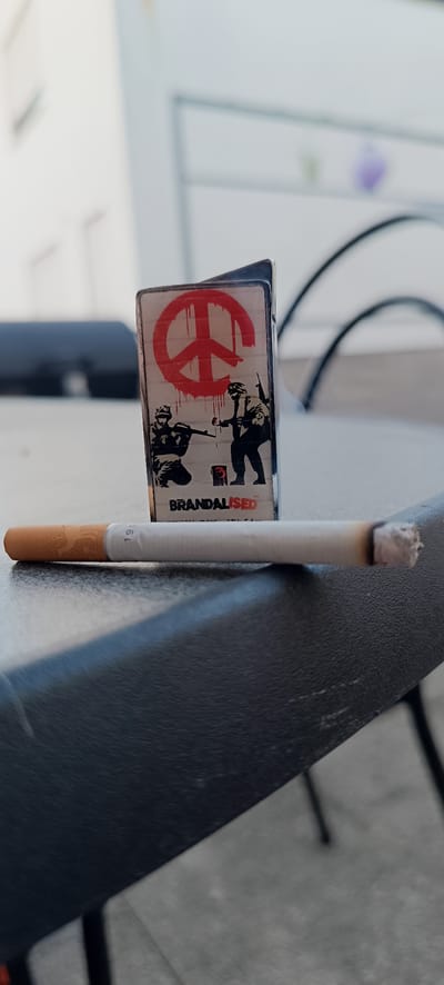 A close-up, low-angle shot shows a lit cigarette lying horizontally across a dark, textured surface, likely a table. The cigarette is positioned in the foreground, with its ash end slightly to the right and showing signs of having been smoked. Behind the cigarette, a silver lighter with a "BRANDALISED" design is standing upright. The design on the lighter features a red peace sign with a dripping effect, and silhouetted figures of two soldiers, one kneeling with a rifle and the other standing, seemingly painting the peace sign on a brick wall. The background is blurred, showing what appears to be an outdoor setting with architectural elements and dark curved shapes, possibly chairs. The scene is illuminated by natural light. The specific location in the city of Winnenden, Germany cannot be determined from the image.