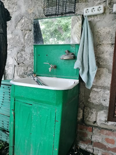 An outdoor sink in Lykhove, Ukraine features a bright green metal cabinet and basin with a silver faucet. A light blue towel hangs on the right side of the sink, adjacent to a triple electrical outlet mounted on a stone wall. Above the sink, a small mirror reflects trees and a dark tree trunk. A miniature lion figurine sits to the left of the mirror. The wall is constructed of rough-hewn stones and mortar, showing signs of age and wear. To the left of the sink, a stack of green wooden planks is visible. The ground below is littered with debris, including what appears to be cigarette butts. The scene appears to be in a weathered, possibly neglected outdoor setting.