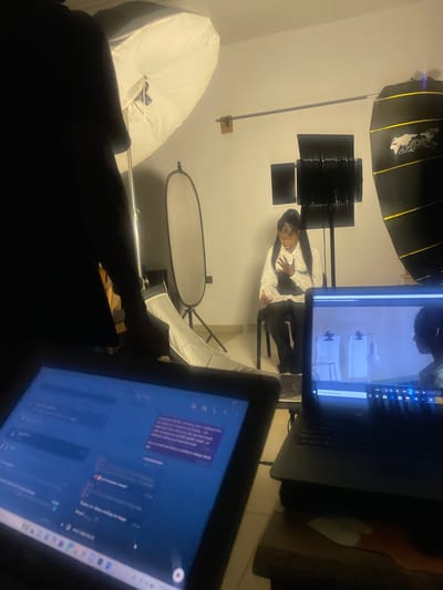 The image depicts an indoor studio setup, possibly for a video shoot or interview, in South Ropp, Nigeria. In the foreground, a dark-clad figure is partially visible, silhouetted against studio lighting. In front of this figure are two electronic devices: a tablet or monitor displaying a chat interface and a laptop.
The central subject of the image is a young Black woman seated on a dark chair, facing slightly to her left. She is wearing a white collared shirt, a black corset or vest, and dark pants. She has her hair pulled back in a high ponytail and is wearing jewelry, including a prominent pendant necklace. Her right hand is on her chest, and her left hand is extended outwards.
The environment is a studio with professional lighting equipment. Two large softbox lights are visible, one to the left and another to the right, illuminating the scene. A smaller, circular reflector is also present. In the background, several monitors are mounted on stands, and a large, dark cylindrical object with yellow stripes is visible on the right. The walls appear to be light-colored.
The laptop in the foreground displays a video feed of the woman on a split screen, suggesting a live monitoring setup. The tablet screen shows a chat conversation with text messages. The overall impression is that of a controlled creative production environment.