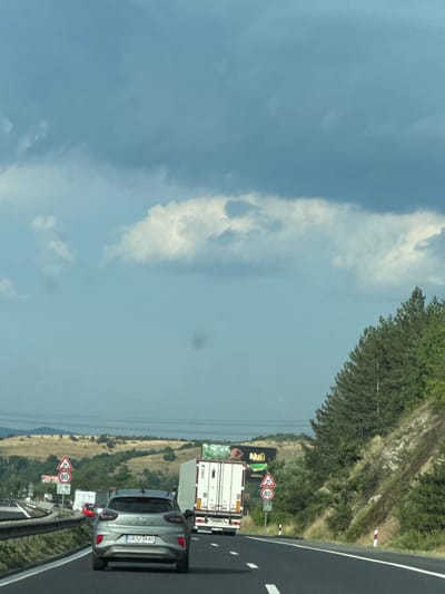 The media file depicts a roadside scene, likely a highway in Dolno Varshilo, Bulgaria. The image is oriented vertically, with the sky occupying the left side and the road and surrounding landscape on the right. The sky is a pale blue with scattered, wispy white clouds, suggesting daytime.

On the right side of the image, a paved road with white lane markings is visible, indicating a highway. A silver car is stopped on the road, and behind it, a white truck is also stationary. Further ahead, other vehicles are lined up, suggesting a traffic jam or a stopped convoy. The landscape beside the road is characterized by a grassy, somewhat arid hillside covered in sparse vegetation and trees.

Several traffic signs are present along the roadside. A triangular sign with a red border and a black symbol of a falling rock or landslide is visible. Next to it is a circular sign with a red border and the number "80" in black, indicating a speed limit of 80. A billboard with the word "Nulit" prominently displayed in large, colorful letters is also visible behind some of the vehicles. The license plate of the silver car reads "CB 5734 AO".

The overall impression is a snapshot of a moment on a Bulgarian highway, possibly during a period of traffic congestion under a bright, partially cloudy sky. The scene appears to be set in a rural or semi-rural area, given the natural landscape surrounding the road.