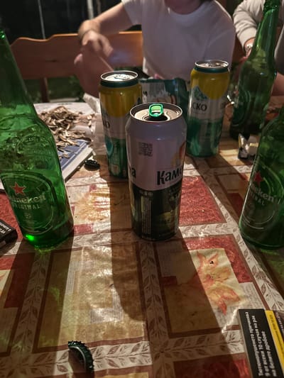 The image depicts a cluttered tabletop scene. Several green glass Heineken bottles and beer cans, including "Каме" (Kame) and "Пиринско" (Pirinsko) brands, are visible. A partial view of a person wearing a white t-shirt is included, showing their arm and hand holding a phone or device. The person is seated at a table. There's a small dish with dried fish and a beer bottle cap on the table surface. The tabletop has a floral-patterned cloth. A black box with text in Bulgarian related to smoking risks is situated on the table as well. The scene suggests a gathering in Voiniagovo, Bulgaria, based on the Bulgarian text on the beer can.