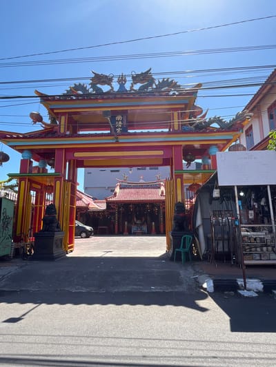 A front-facing, eye-level shot captures a vibrant, ornate Chinese temple gate with a clear blue sky above. The gate is predominantly red and yellow, adorned with two dragon sculptures on its roof and traditional Chinese characters above the archway. To the left of the gate, a black stone lion statue stands guard. Beyond the gate, the temple complex is visible, featuring red-tiled roofs, lanterns, and more intricate architectural details. A silver car is parked inside the temple grounds. In the foreground, the asphalt road is visible, with shadows cast by overhead power lines and a streetlamp. To the right of the gate, a makeshift stall covered with a gray tarp displays various items, and a green plastic chair sits near the temple entrance. The environment appears to be a street in Desa Tambakreja, Indonesia, under bright sunlight.