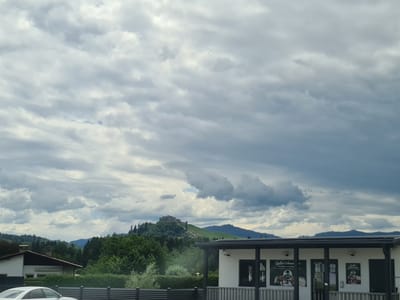 The image depicts a cloudy sky over a landscape with a building in the foreground and a castle on a hill in the background. The building in the foreground is a modern, single-story structure with a black roof and white walls. It appears to be a commercial establishment, as indicated by signage on the windows. The building has a covered porch area supported by black pillars. In front of the building, a black fence runs horizontally across the frame. To the left of this building, a portion of a house is visible, with a white wall, a dark roof, and a chimney. A white car is parked in front of the fence.
In the middle ground, a densely wooded hill rises, covered in green trees. The castle is situated atop this hill, a prominent structure with stone walls and towers, appearing somewhat ruined but still substantial. A path or road is visible winding up the side of the hill towards the castle.
The background consists of rolling hills and mountains, covered in trees, fading into the distance under a heavy, overcast sky with large, dramatic clouds. The overall scene suggests a rural or semi-rural setting in or near the city of Frauenstein, Austria, given the presence of the castle, a common feature of the Austrian landscape. The lighting is diffused due to the cloudy conditions.