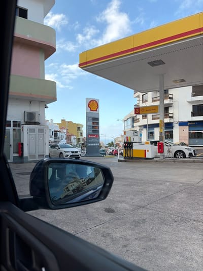 The image displays a bright, daytime scene at a Shell gas station, viewed from inside a vehicle. The primary image is rotated 90 degrees counter-clockwise; describing it as if correctly oriented, the ground would be horizontal.

In the foreground, part of a car's interior, likely the dashboard or door frame, is visible along the bottom and right edges of the frame. A black side mirror is prominent on the right side, reflecting a distorted but recognizable view of the gas station canopy, surrounding buildings, and potentially a person's hand or arm inside the car.

The main subject is a Shell gas station. A large, flat canopy, painted in Shell's characteristic yellow with a red stripe, extends across the upper left and central parts of the frame, providing shade over the fueling area. Underneath this canopy, several fuel pumps are visible, also adorned with yellow and red Shell branding. A small sign attached to a support pillar under the canopy reads "Quality Fuels" and features a red square with the number "3" inside.

To the right of the canopy, a tall, vertical Shell sign with the iconic red and yellow scallop logo is clearly visible. Below the logo, fuel prices are displayed digitally. While partially obscured, prices like "1.18" and "2.99" are discernible, along with the text "V-Power," indicating a premium fuel type.

The ground of the gas station is a paved, grey concrete or asphalt surface. Several other vehicles, including a white car, are visible parked or waiting near the pumps and in the background.

Behind the gas station, various multi-story buildings line a street. Some buildings have light-colored facades, and one in the mid-left features distinctive light pink and light green curved architectural elements. Further back, more buildings are visible, suggesting an urban or suburban setting. The sky above is a clear, light blue with scattered white clouds, indicating fair weather. The overall impression is a bustling gas station during daylight hours.