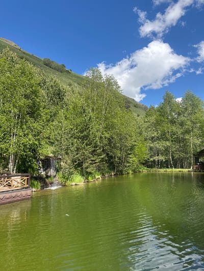 The image shows a serene lake with green water, reflecting the blue sky and white clouds. A small waterfall cascades into the lake from a rocky structure surrounded by lush green trees. On the left side of the lake, a wooden deck with a railing is visible, extending over the water. The background is dominated by a steep, grassy mountain slope covered in trees, typical of the natural landscape around Terskol, Russia. The scene is tranquil and idyllic, suggesting a natural resort or park setting in the Elbrus region.