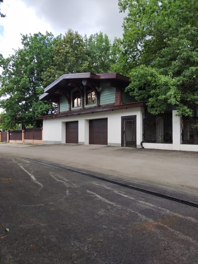 The image depicts a two-story house with a distinctive architectural style, likely a garage attached to the main residence. The lower level features two dark brown garage doors, set within a clean white facade. Above the garage, the second story is clad in a pale green material and features a steeply pitched, dark brown roof with exposed wooden beams, creating a rustic yet modern aesthetic. Two large windows on this upper level reflect the surrounding trees and cloudy sky.
The house is situated behind a white fence and a decorative wrought-iron gate, hinting at a private property. To the left, a wooden fence with brick pillars marks the boundary of the property. Mature trees with lush green foliage frame the house, suggesting a setting with abundant nature. A paved driveway with a drainage grate runs in the foreground.
The scene appears to be set in the daytime, under an overcast sky with some clouds visible. The lighting suggests it could be late afternoon or early evening. There are no people visible in the image, and the overall mood is quiet and serene, with no clear activity taking place. The location context of Blokhino, Russia, places this scene in a possibly suburban or rural setting within the country. There is no visible text in the image.