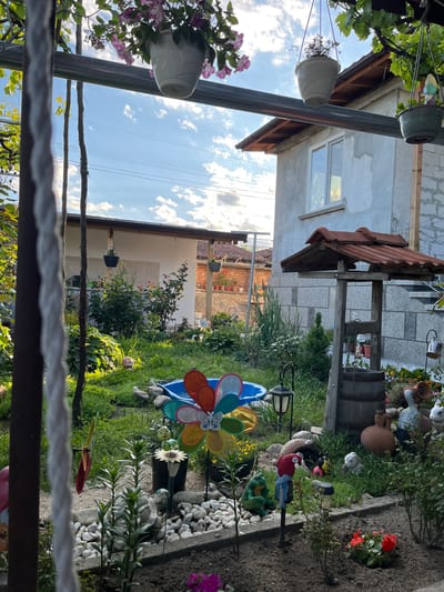 The image depicts a backyard garden scene in Karavelovo, Bulgaria, viewed from a partially obstructed perspective. A variety of garden decorations are present, including pinwheels, flower pots, and ceramic figures amongst foliage and white gravel paths. The architecture visible includes portions of residences with tiled roofs and plastered walls. Overhead, a pergola structure with hanging planters is partially in view. The sky is blue with scattered clouds.