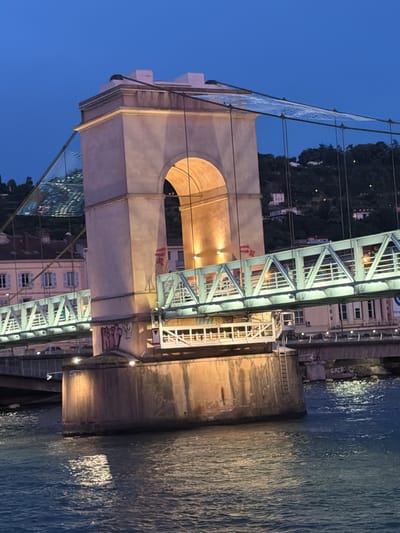 This is a vertical, slightly tilted, night-time photograph of a bridge over a body of water, likely a river, in Vienne, France. The bridge features a large, light-colored stone tower with an arched opening illuminated from within. Above the tower, a metal truss structure of the bridge is visible, lit by warm lights. The bridge appears to be part of a larger infrastructure, with railings and cables supporting it.

To the left of the bridge, a hillside covered with trees and some buildings is visible, also illuminated by lights. Reflected in the water are the lights from the bridge and the surrounding buildings, creating shimmering patterns. The sky is a deep blue, indicating it's dusk or early night. The overall mood is serene and picturesque, highlighting the architectural and natural beauty of the scene at night.

There are some instances of visible text in the image:
- Red graffiti, possibly stylized letters, on the side of the metal truss structure.
- White lettering on a red sign on the bridge structure.
- The word "GRZA" spray-painted in white on a concrete wall near the water's edge.
- Possibly some Chinese characters on a sign on a building in the background, though this is not clearly legible.