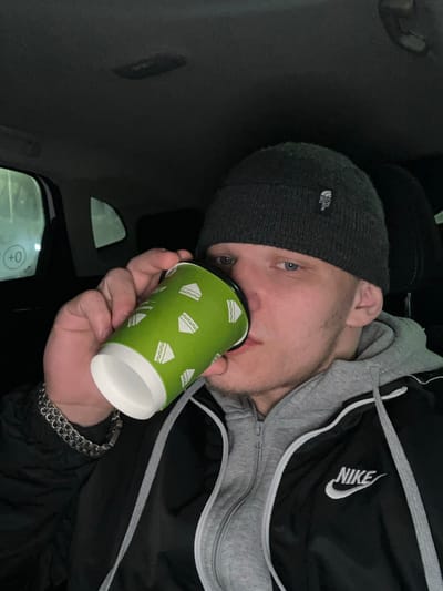 The image displays a young adult male seated inside a vehicle in Dobryanka, Russia. The subject has light skin and blue eyes. He wears a dark grey knitted beanie with a white "The North Face" logo patch, a grey hooded sweatshirt, and a black jacket bearing a white Nike logo on the left chest. A silver-colored chain link bracelet is present on his left wrist. He is actively drinking from a green paper cup, decorated with white sandwich icons, holding it to his lips with his right hand. The vehicle's interior is dimly lit, and a numerical display showing "+0" is visible through a rear passenger window on the left.