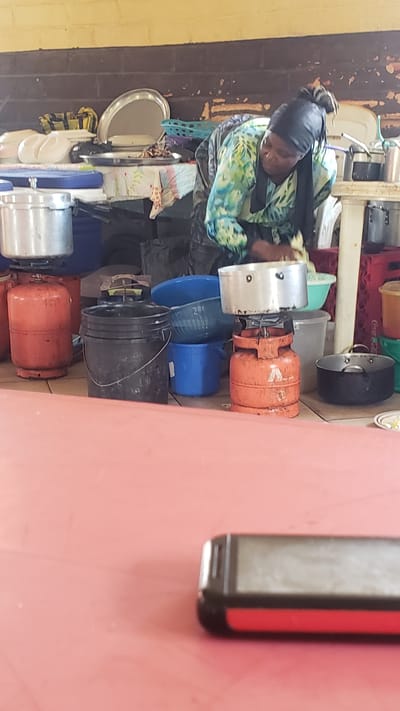 In Unguwar Shanu, Nigeria, a woman wearing a headscarf and a patterned long-sleeved shirt is engaged in cooking outdoors. She is crouched beside a portable gas stove with a pot on top, appearing to be washing or cleaning something. Surrounding her is a collection of kitchenware and cooking supplies, including large metal pots, plastic buckets of various sizes and colors (black, blue), and a blue colander. Two orange gas cylinders are prominently visible, one directly beneath a pot and another to the left. The setting appears to be an open-air or semi-enclosed area, possibly a market or an outdoor kitchen, with a brick wall and a light-colored ceiling overhead. In the foreground, a pink surface is visible, with the corner of a black and red electronic device, likely a mobile phone, resting on it.