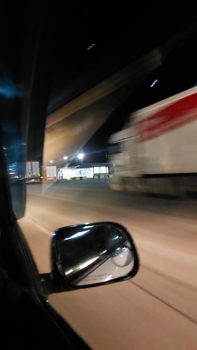 The image captures a blurry nighttime view from inside a vehicle, likely in motion, looking out the passenger side. A side-view mirror is visible in the foreground, reflecting the road and some overhead lights. Beyond the mirror, a road with multiple lanes is visible, illuminated by streetlights. Two semi-trucks are present on the road; one is stationary on the left side, and another is in motion on the right, with its red and white markings blurred by speed. In the background, a building with large windows and bright lights appears to be a commercial or industrial facility. The overall scene suggests a highway or a large parking area at night. The image is too blurry to ascertain any specific details about the city of Дербісек ауылдық округі, Kazakhstan, or any individuals present.