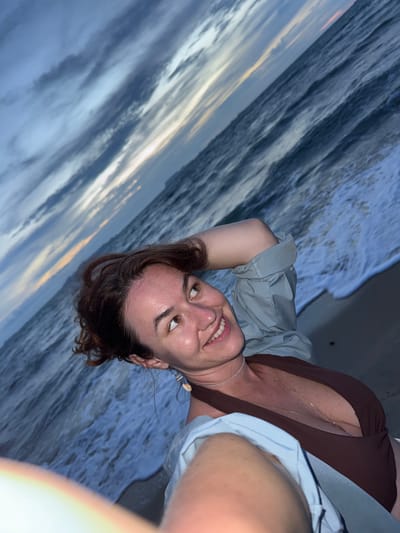 A woman with reddish-brown hair is standing on a beach at dusk, looking up and to her left with a smile. She has on a brown swimsuit and a light blue shirt. The ocean is behind her, with waves lapping at the shore. The sky is cloudy, with streaks of orange and yellow from the setting sun visible. The image is taken from a low angle, with the woman's arm in the foreground. The scene appears to be located in the city of Tong Yang, Thailand.