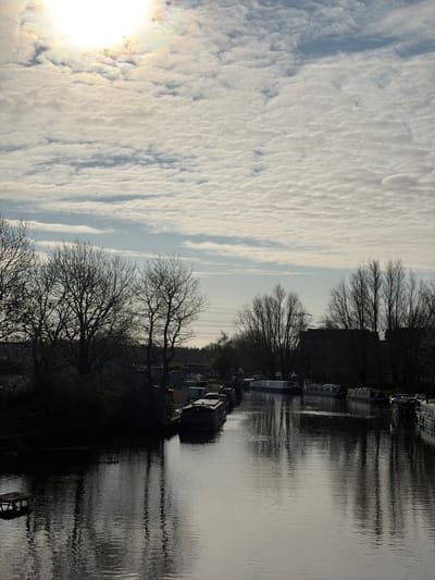 The image depicts a static scene along a calm waterway in Hoddesdon, United Kingdom, likely a section of the River Lea Navigation. The perspective is from an elevated position looking down the length of the water.

The upper third of the frame shows a bright, partially cloud-covered sky. The sun is visible in the upper left, appearing as a strong, bright light source with a slight iridescent halo caused by atmospheric conditions. The clouds are predominantly scattered altocumulus or similar formations.

The central and lower portions of the image are dominated by the waterway, which reflects the sky and the surrounding silhouettes. Both banks are lined with numerous deciduous trees, bare of leaves, indicating either winter or early spring. These trees cast dark, stark silhouettes against the brighter background, with their reflections visible in the water.

Along both banks, multiple narrowboats or similar leisure craft are moored. These vessels are generally dark in color and appear to be unoccupied. On the left bank, partially obscured by the trees, there are hints of structures or facilities, possibly a marina or maintenance area. A small, dark wooden platform or pier extends into the water from the left bank in the foreground. On the right bank, behind the trees, distant dark-roofed buildings, possibly industrial or residential, are visible. Overhead power lines or pylons are faintly discernible above the treeline in the mid-right background.

No individuals are present in the visible scene. The water surface is smooth with minimal ripples, suggesting calm conditions. The overall lighting suggests a bright, possibly overcast day, with the sun positioned to create strong backlighting.