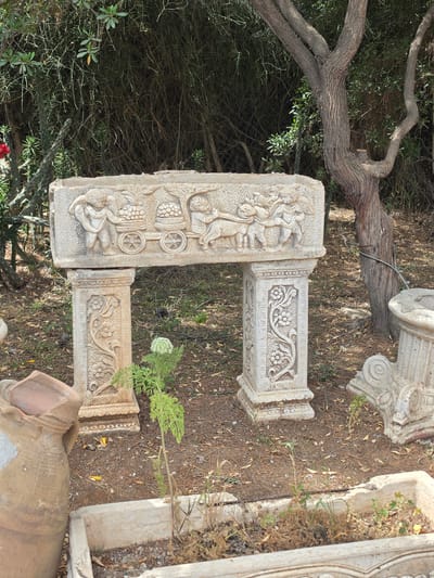 The image shows a collection of weathered, decorative stone objects in an outdoor setting, likely in Mate Yehuda Regional Council, Israel, suggested by the landscape characteristics. The objects consist of sculpted stone blocks with intricate carvings, possibly fragments of architectural structures. The carvings depict figures and floral designs, indicating an origin from a decorative or artistic context. The stones are placed among dry leaves, scattered vegetation, and near a large tree branch, possibly in a garden or an area with neglected artifacts. The presence of additional fragments and a clay vessel suggests the location is a storage area for discarded architectural pieces. There are no people in the scene.