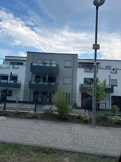 The image shows a modern apartment building in Volpertshausen, Germany, with a cloudy sky above. The building features several balconies with dark railings and some greenery on them. A paved walkway runs in front of the building, separated by a black metal fence and a strip of landscaping with small trees and bushes. A lamppost with a sign is visible to the right of the building. The architecture is contemporary, with clean lines and a mix of white and grey facades. The overall impression is that of a well-maintained residential area.