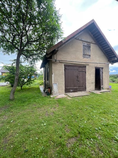 This image depicts a rustic, weathered barn or outbuilding in a rural setting, likely within the Бучацінскі сельскі Савет of Belarus. The structure is characterized by its rough, beige plastered walls and a steeply pitched, rust-colored metal roof. A wooden window with shutters is visible on the upper facade. The main entrance features large, dark brown double doors with visible hardware and a padlock. To the right of these doors is a single, smaller wooden door, partially ajar, revealing darkness within. Both entrances are fronted by wooden planks serving as makeshift walkways over the grassy ground.
The barn is situated on a patch of uneven, green lawn, with scattered leaves and patches of bare earth. To the left of the barn, a mature tree with a thick trunk and leafy branches casts some shade. In the background, other rural dwellings and vegetation are visible, suggesting a residential area. The sky above is overcast with some white clouds. Various small objects, including pots and a discarded window frame, are scattered near the side of the barn. The overall impression is one of an aging, functional outbuilding in a quiet, rural Belarusian landscape.