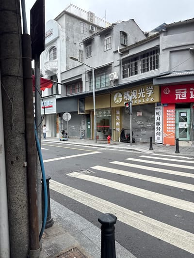 The image depicts a street scene in Liwan District, China, likely in the daytime with overcast skies. A pedestrian crossing with white stripes is prominent in the foreground, leading towards a sidewalk lined with bollards and electrical poles. Various shopfronts are visible on the right side of the street, displaying signs in Chinese. One shop has a yellow facade with Chinese characters and a red traffic light above it. Adjacent to it, a red shopfront displays more signage. The buildings appear to be of multiple stories, with windows featuring metal grilles. A black scooter is parked near the red traffic light. On the left side of the image, a utility pole with wires is partially visible, along with a blue hose coiled around it. A person in a light-colored shirt and dark pants stands on the sidewalk near a shop entrance. The overall impression is of a commercial street with some businesses closed, indicated by the roll-down shutters on some storefronts.