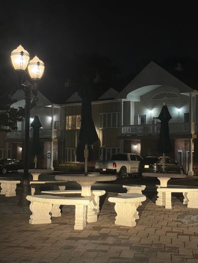The image shows a nighttime scene in the Village of Amityville, United States of America. In the foreground, there are several ornate white stone benches and tables arranged on a brick-paved area. Two tall, decorative lampposts with illuminated globes stand to the left, casting a warm light. In the middle ground, two large, dark green umbrella-like structures are visible, likely protecting the seating areas. Beyond these, a row of buildings with balconies and illuminated windows suggests residential or commercial units. Several vehicles, including a white pickup truck and a dark sedan, are parked in front of the buildings. The sky is dark, indicating it is nighttime.