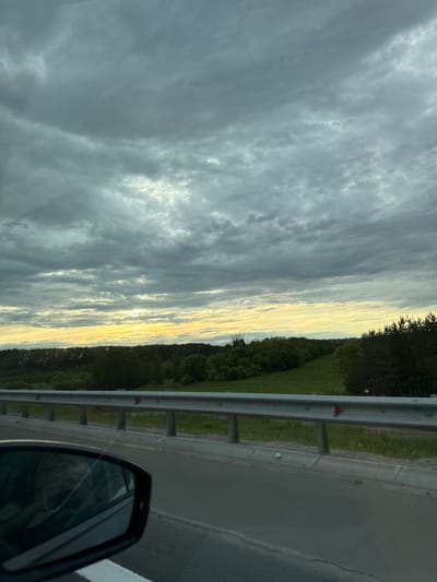 The image captures a roadside view during what appears to be a cloudy sunset or sunrise. The dominant subjects are the sky, a roadside guardrail, and a paved road. The sky is filled with a mix of dark grey and lighter, illuminated clouds, suggesting dynamic weather conditions. A sliver of bright light, likely from the sun, peeks through the clouds near the horizon, casting a warm glow on the sky.
To the right of the guardrail, a landscape of dense green trees and fields is visible, stretching into the distance. The guardrail itself is made of metal and runs parallel to the road. The road, which takes up the right side of the frame, is asphalt and has a white line marking its edge. A portion of a car's side mirror is visible in the bottom right corner, indicating the image was taken from inside a vehicle.
The overall scene conveys a sense of travel or journey. The time of day is suggested by the light quality in the sky, likely late evening or early morning. The weather is overcast but with potential for clearing or having recently cleared. No people or explicit activities are visible, but the context of being on a road implies movement. There is no visible text in the image.
