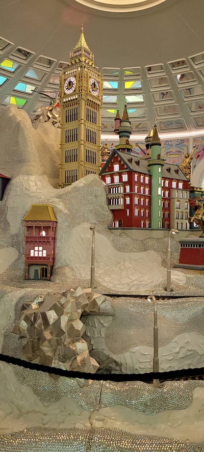 The image displays a detailed model of a city, featuring several buildings and a prominent clock tower reminiscent of Big Ben. The structures are intricately designed, with bright red and green accents on some buildings, and a golden spire on the clock tower. The city is set against a backdrop of textured, mountainous terrain, covered in what appears to be small, reflective embellishments, giving it a shimmering, almost crystalline appearance. Streetlights are strategically placed throughout the model. The overall scene is enclosed by a curved ceiling with a grid of illuminated panels, suggesting an indoor display or exhibition. This model is located within the Swarovski Kristallwelten (Crystal Worlds) in Wattens, Austria, a popular tourist attraction known for its art installations and crystal creations. There are no people visible in the image.