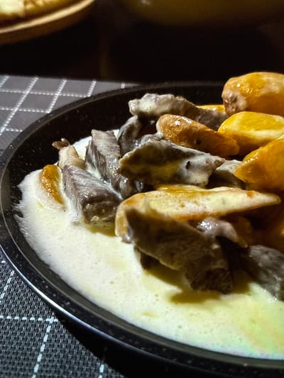This is a close-up, eye-level, indoor shot of a dish of food. The food appears to be beef stew with potatoes or dumplings in a creamy sauce. The dish is black and round, and it sits on a dark grey and white checkered placemat. The background is dark and out of focus, making it impossible to determine the specific location within the city of Artik, Armenia. There are no people or animals visible in the image. The lighting is dim, with a slight yellow cast.