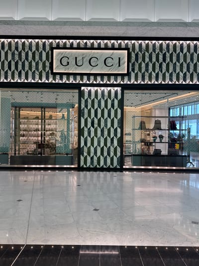 A Gucci storefront is depicted in a brightly lit indoor commercial area, likely an airport terminal or shopping mall, in Roissy-en-France, France. The store's facade features a prominent central illuminated "GUCCI" sign with black lettering on a white marble-like background, framed by black. This sign is set against a geometric pattern of alternating dark green/black and white/light gray parallelogram-shaped tiles, arranged vertically to create a three-dimensional effect. This tile pattern also covers the central column between the two main display windows.
The store has two large glass display windows. The left window showcases multiple shelves containing various footwear, predominantly shoes and boots, and potentially some small accessories or bags, all illuminated from within. A faint reflection of the word "PRADA" is visible on the glass of this window. The right window displays several handbags, backpacks, and hats on multiple shelving units. The store's interior beyond the displays appears spacious and well-lit with recessed lighting.
The floor in front of the store consists of polished light-colored marble or stone tiles with subtle gray veining and occasional darker square inlays. Closer to the viewer, a strip of polished dark, possibly black, tiles runs horizontally, featuring small embedded lights creating a reflective sheen. The overall scene indicates a clean, upscale commercial environment. No individuals are visible in the image, nor are any actions or interactions occurring.