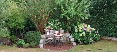 This is a medium shot, eye-level, full-frame, wide-angle photograph of a small, secluded garden seating area. The scene is dominated by lush green foliage, with a variety of trees and flowering bushes. In the center of the image, a circular brick patio is partially enclosed by a low, segmented stone wall. On the patio, a small, round table with a dark, possibly metal, base and a lighter colored top is set with two plates and a small, dark object that resembles a cake or a pie. Two dark metal chairs with decorative backs are placed at the table. Behind the table, on the stone wall, there are two decorative plates. To the left of the seating area, a neatly trimmed, rounded bush stands out against the taller trees. To the right, a large flowering shrub with clusters of pale green and pink blooms is prominent. The background is densely packed with trees and shrubs, suggesting a private garden setting. The lighting indicates it is daytime, likely overcast or late in the day, as there are no harsh shadows. The image is slightly soft focus, contributing to a painterly quality. The setting is consistent with the typical appearance of gardens in the Bavarian town of Garching an der Alz, Germany, known for its verdant landscapes and well-maintained private outdoor spaces.