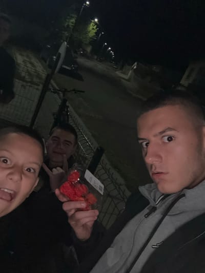 Three young men are visible in a dark outdoor environment. The man closest to the camera has short hair and an open mouth. Another man in the mid-ground is making a hand gesture. The third man, in a grey hooded jacket, is holding a container of red fruits, possibly raspberries or strawberries. Fencing is in the background. Streetlights and a building are visible in the distance. The location is potentially in the city of Goritsa, Bulgaria.