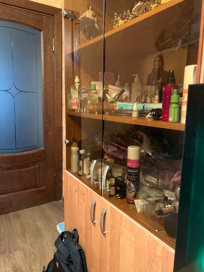 The image shows a close-up of a wooden cabinet with glass doors, filled with various items. The cabinet is located indoors, near a wooden door and on a wooden floor. On the top shelf, there are small figurines and a framed picture of a woman. The shelves below are filled with bottles, cans, and other personal care products. A black backpack is visible on the floor in front of the cabinet. The environment appears to be domestic. This scene is not located in Tula, Russia.