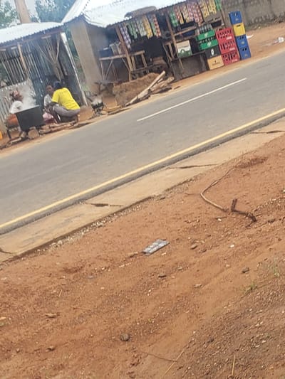 In Gumel, Nigeria, an outdoor food stall is situated next to a paved road with a yellow dividing line. Several individuals are present, with two actively engaged in preparing food. One person, wearing a bright yellow shirt and grey trousers, is crouched near a steaming pot. Another person, partially obscured, is also near the cooking area. The stall itself is constructed from corrugated metal and wood, displaying various goods on shelves, including rows of bottled beverages in colorful crates. The surrounding environment is earthy and arid, with scattered debris on the ground.