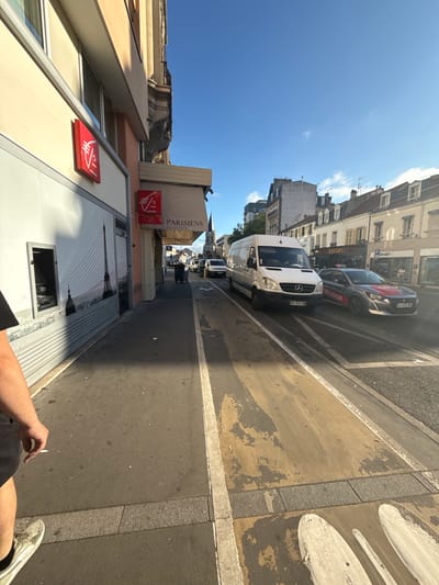 The image depicts a street scene in Colombes, France, under a clear blue sky. On the left, a building facade features a red square sign with a white logo, likely representing a bank, and a large mural of the Eiffel Tower. A person's legs and arm are visible in the foreground on the left, holding a cigarette.
The street itself has a marked lane for vehicles and a pedestrian walkway. Several vehicles are present: a white van with a Mercedes-Benz logo, a dark gray car with red and white decals, and other cars further down the street. Buildings line the right side of the street, with some displaying signs for businesses such as "ZER GRILL" and "FORÊT". In the background, a church spire is visible. The environment appears to be an urban setting, possibly a commercial or residential street. The lighting suggests it is daytime, with sunlight casting shadows.
