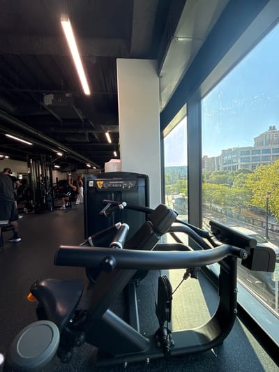 An indoor gym, likely located in the City of Yonkers, United States of America, is depicted from a low-angle, close-up perspective on a weight machine. The machine, a black shoulder press with a dark gray seat and orange adjustment lever, occupies the foreground, casting a shadow on the black rubber floor. Sunlight streams through a large window to the right, illuminating the gym equipment and the exterior scene. Beyond the window, a tree-lined street with a river, buildings, and a clear sky are visible. In the background of the gym, other weight machines and a few individuals, including a man in black shorts and a woman on a lat pulldown machine, are present. Fluorescent lights are installed on the dark ceiling, and exposed ventilation ducts are also visible.