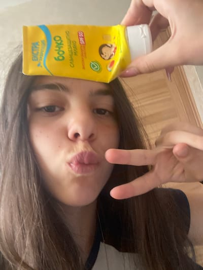 A young woman is pictured holding a yellow bottle labeled "BONKO Слънцезащитно мляко SPF 50 ЕКСТРА." She is making a peace sign with her other hand and pursing her lips. Her hair is dark brown and wavy. The background is out of focus and appears to be an interior space with wooden features. There is no evidence to suggest that the location is Dragovishtitsa, Bulgaria.