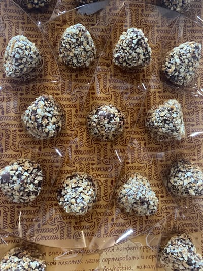 The image shows a tray of chocolate candies covered in chopped nuts. The candies are arranged in individual compartments within a transparent plastic tray, which rests on a patterned surface. The pattern consists of the repeated word "Россия" (Russia) in a cursive font. The candies vary slightly in shape, some being rounded and others more angular or heart-shaped. The chopped nuts cover most of the chocolate surface. Text is visible on the right side of the image, indicating the tray is made for candy sorting. The setting is indoors, with soft lighting.