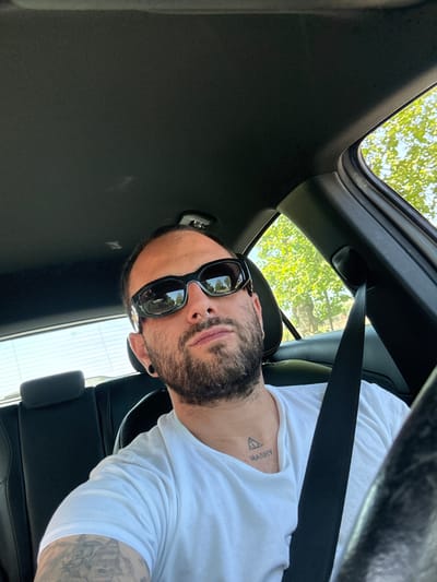 A bearded man with sunglasses and a tattoo on his neck is wearing a white t-shirt and a seatbelt while sitting inside a car, likely a modern model, with a sunroof. The car's interior is dark. Outside, trees and sky are visible through the sunroof and a side window. The exact location in Nettuno, Italy, cannot be determined from the image.