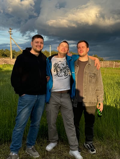 Three men are lying on the grass in Vysokinichi, Russia, facing the camera. The man at the top is holding a green bottle and has his arm around the man below him. All three men are fully clothed. The background includes a fence, electrical poles, and a partly cloudy sky.