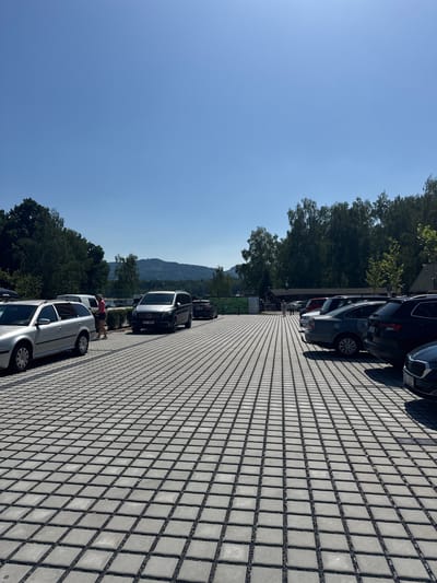 This image depicts a paved parking lot with a hexagonal tile pattern, possibly made of concrete pavers. The lot is lined with several cars, including a silver station wagon in the foreground to the left, a dark-colored van with license plate "6L7 6858" in the middle, and a dark SUV on the right. A woman in a pink shirt and dark shorts is standing near the silver station wagon, looking at her phone. In the background, there are trees and hills under a clear blue sky, suggesting an outdoor, possibly rural or suburban setting. The scene appears to be in Hrádek nad Nisou, Czechia, given the context provided. The lighting indicates it is daytime, with the sun casting shadows.