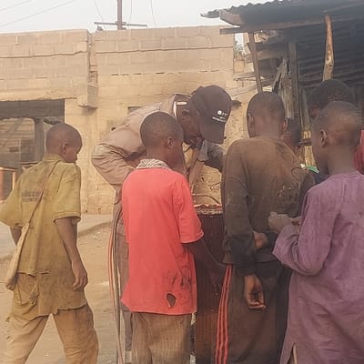 An outdoor scene in Gudi, Nigeria, shows an adult male and five children gathered around a large, metallic object, likely a vehicle radiator, under diffuse daylight conditions. The adult, positioned centrally and wearing light-colored coveralls and a dark baseball cap with a logo, is bent over, focused on the radiator, with what appears to be a hose or pipe near his hands. Five young boys encircle the adult, closely observing his activity. Their clothing, consisting of various shirts and pants, is noticeably soiled and exhibits signs of wear and tearing; one child wears a yellow-green shirt with text on the back, another a red torn shirt, and a third a dark brown shirt with a faded graphic on the back. The background features an unfinished or partially damaged building constructed from unpainted concrete blocks to the left, and a more rustic structure with a corrugated metal roof supported by wooden posts to the right. Overhead, utility lines and a pole are visible against a hazy sky. The ground surface is compacted dirt.