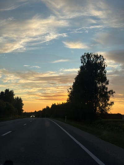 This image depicts a rural road in Aizupes, Latvia, during a sunset. The sky is filled with wispy clouds streaked with orange, yellow, and blue hues. A large, dark silhouette of a tree stands prominently on the right side of the frame. The road itself is asphalt with white lane markings and stretches into the distance, where a single car with its headlights on is approaching. The surrounding landscape is characterized by dark silhouettes of trees and vegetation, suggesting a wooded area or fields bordering the road. The overall atmosphere is serene and atmospheric, indicative of the twilight hours.
